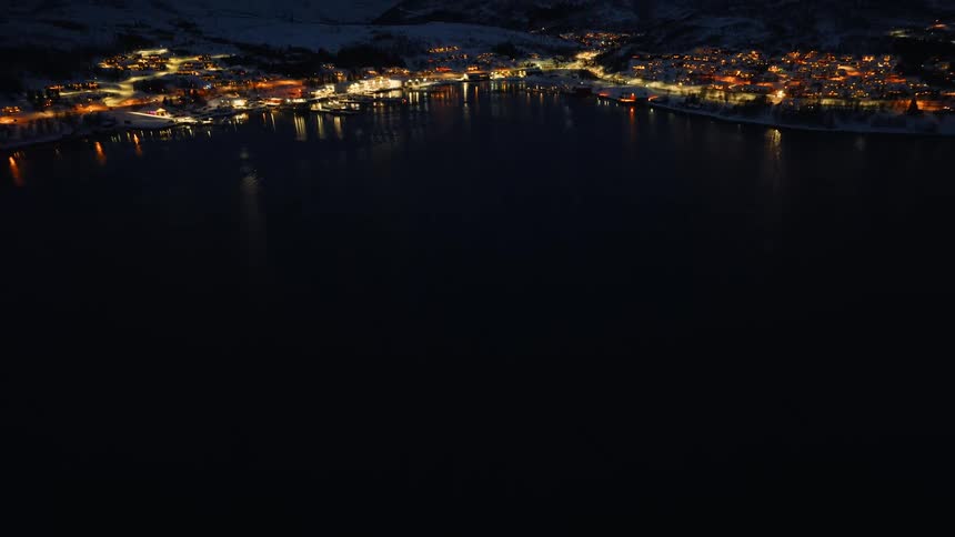 A slow aerial tilt-up reveals the illuminated coastal town of Skjervoy, Norway, surrounded by a dark fjord and snowy mountains at night