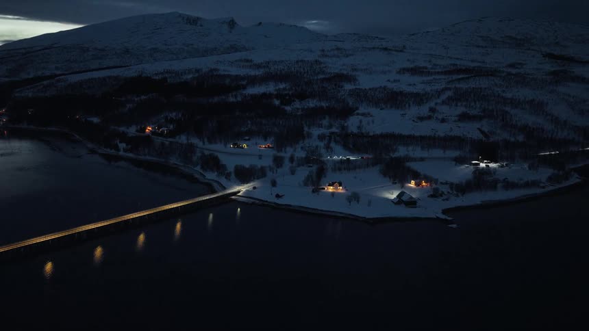 A tranquil aerial view shows an illuminated bridge crossing dark waters to a small, snow-covered coastal village at dusk