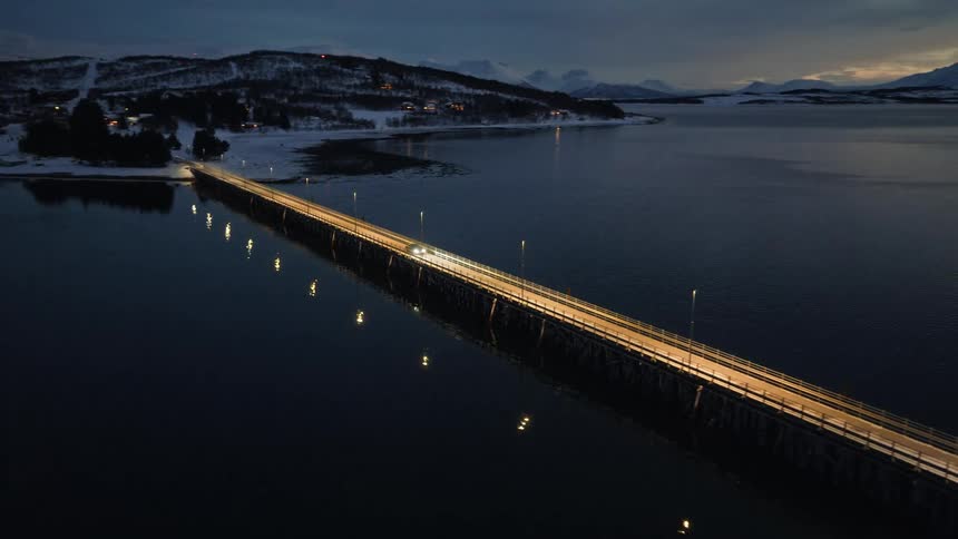 Serene aerial view of a car driving across a long, illuminated bridge over a fjord at dusk, surrounded by snow-covered mountains