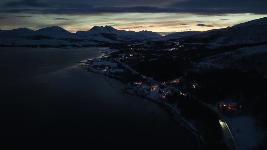 A beautiful aerial view of a snowy coastal village in the mountains at twilight as cars drive along the shore