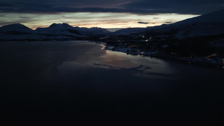 An aerial view captures a tranquil fjord at twilight, with the sunset reflecting on the water next to a snowy mountain village