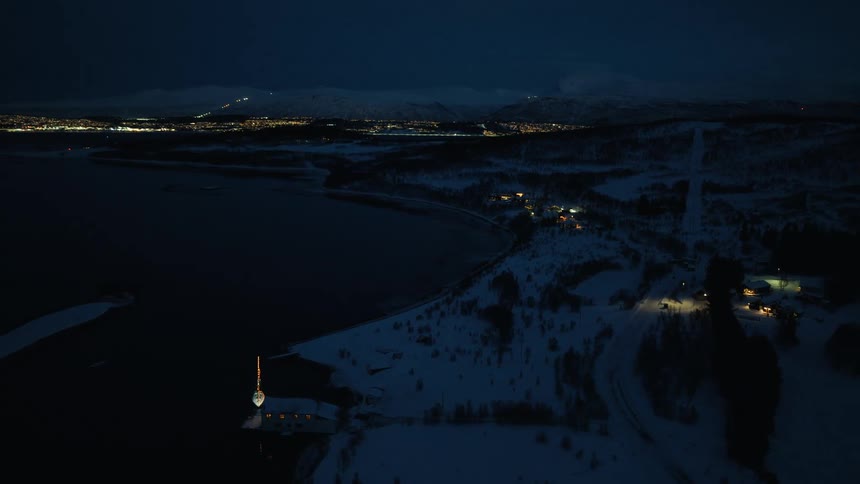 Expansive aerial view of a snow-covered coastline at night, with a glowing town visible across the dark water under a twilight sky