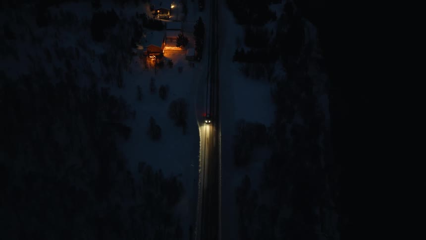 An aerial top-down view follows a car with bright headlights driving along a remote, snow-covered road at night