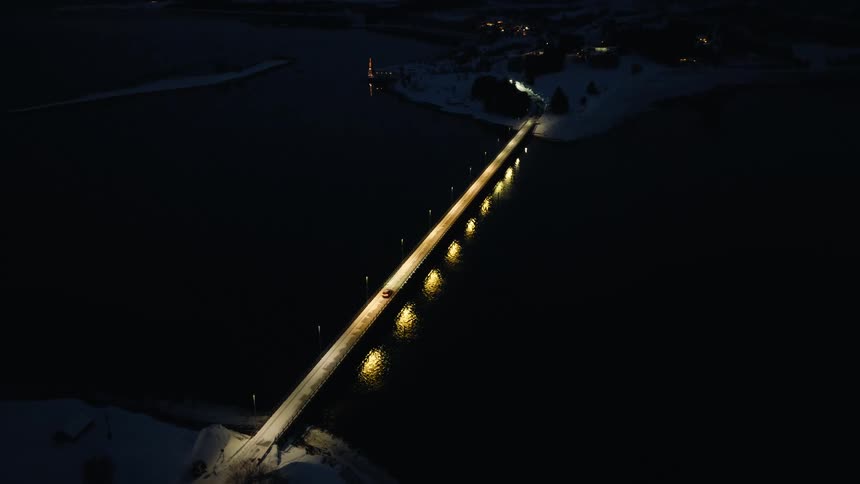 High angle aerial shot follows cars driving over a long, illuminated bridge spanning dark water, with a snowy landscape and city lights in the distance at night