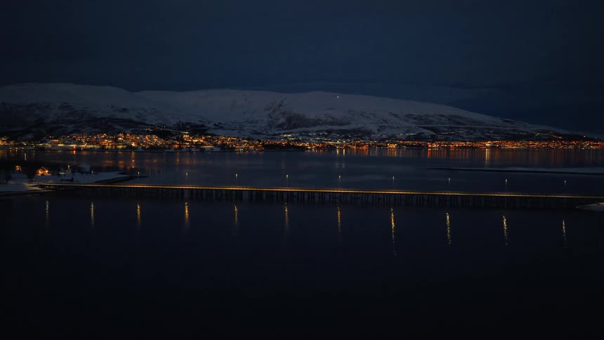 Expansive aerial view of a brightly lit bridge crossing a dark fjord at night, with the glowing lights of a Norwegian town and snowy mountains in the distance