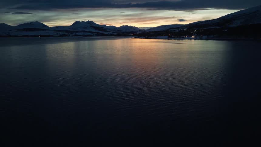 A dramatic aerial shot pulls back from dark rippling water to reveal a vast fjord surrounded by snowy mountains at twilight