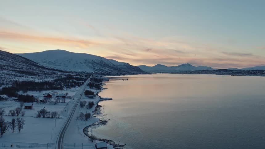 An aerial drone slowly rises over a tranquil, snow-covered Norwegian village on the edge of a fjord at dusk