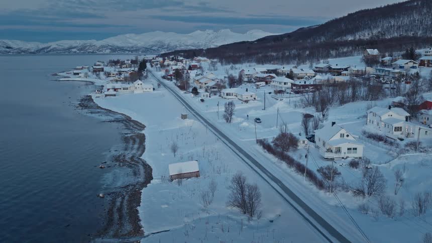 A slow aerial ascent reveals a picturesque snow-covered coastal town nestled beside a calm fjord with mountains in the distance
