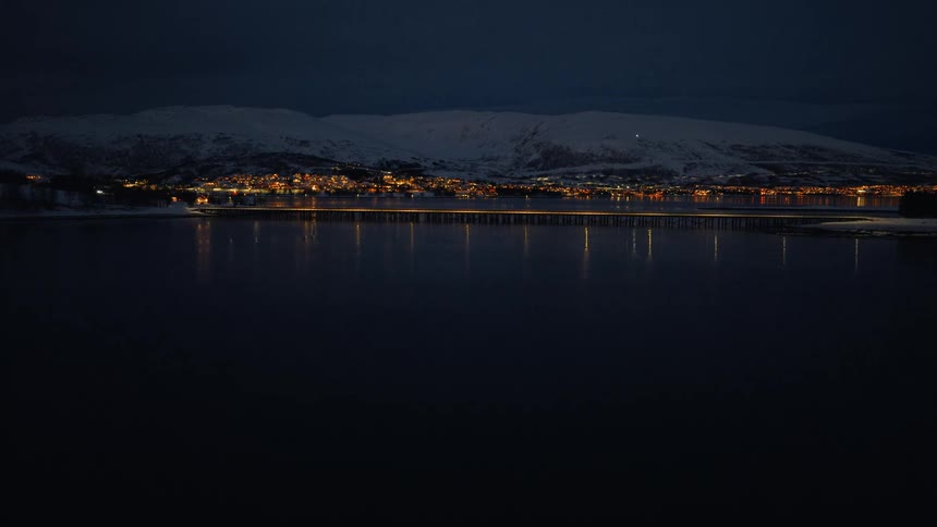 A tranquil aerial view of the glowing city lights of Tromsø and the Tromsø Bridge reflected in the calm fjord at night, with snow-covered mountains in the background