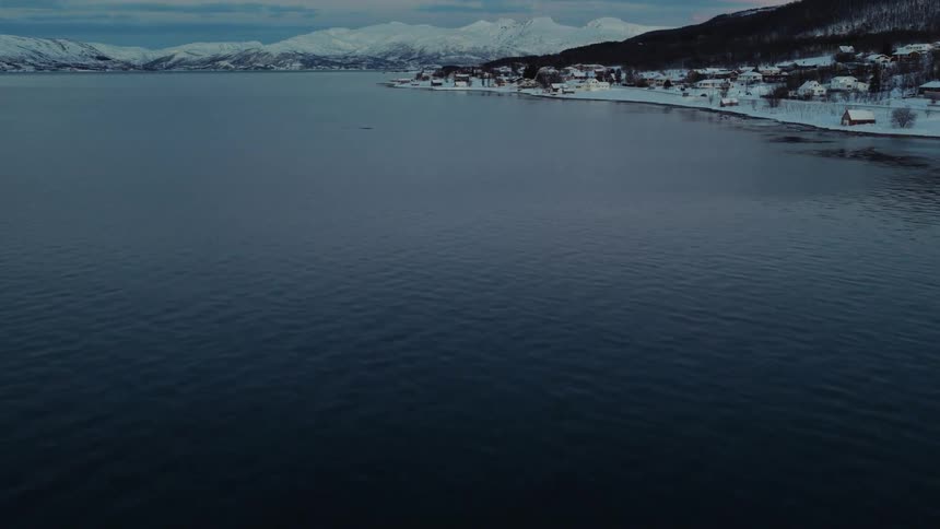 An aerial shot tilts up from the dark, cold water of a fjord to reveal a quiet, snow-covered village and majestic mountains in a winter landscape at dusk