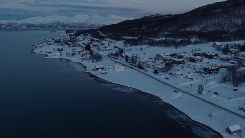 A slow aerial push-in shot over a serene, snow-covered coastal town nestled beside a calm fjord and mountains during a wintery blue hour