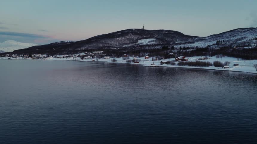 An aerial shot slowly approaches a tranquil, snow-covered village on the shore of a fjord during the winter blue hour