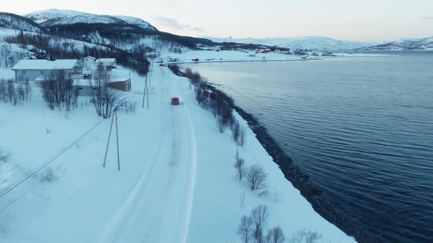 Aerial shot follows a red van driving on a snowy coastal road through a remote Norwegian village in winter
