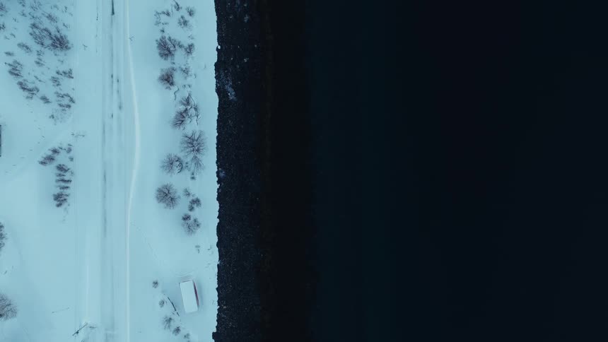 An ascending top-down aerial shot captures the stark contrast between a snow-covered coastline and the dark, tranquil water