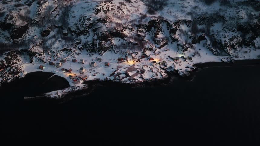 Top-down aerial shot of a cozy, illuminated village on a snowy coastline during a winter twilight