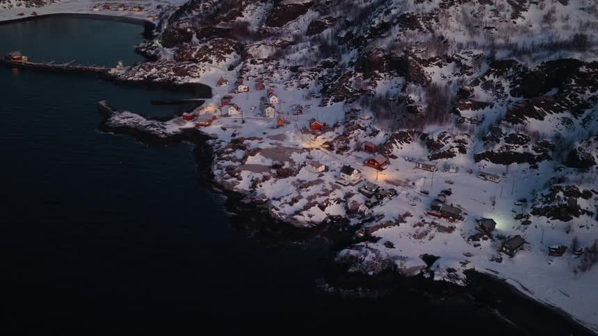 Aerial drone view slowly descends over a quaint, snow-covered fishing village nestled on a rugged coastline at twilight
