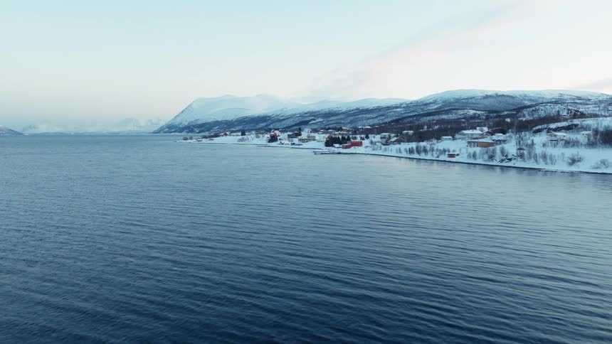 Slow aerial shot over a calm fjord towards a remote, snow-covered village at the base of mountains in a vast winter landscape