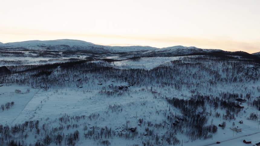 An aerial view captures a lone car driving on a remote road through a vast, snowy Norwegian landscape alongside a fjord at sunset