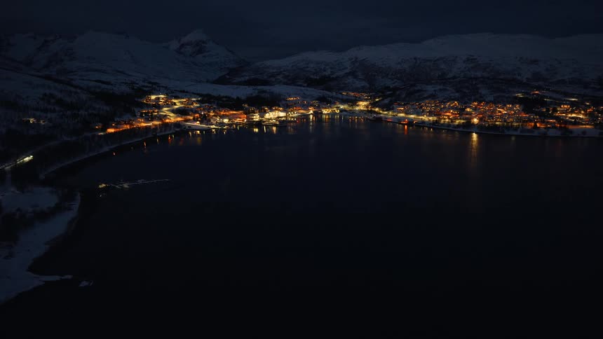 A stunning aerial view shows a cozy, illuminated coastal town nestled by a fjord between snow-covered mountains on a dark winter night
