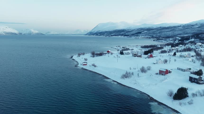 Slow aerial dolly shot over a tranquil, snow-covered coastal village in a Norwegian fjord during a serene winter twilight