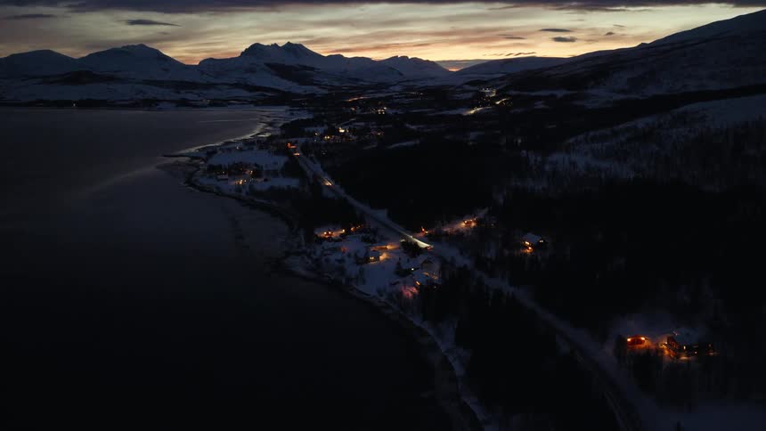 An aerial view shows a snowy coastal town glowing with warm lights as cars drive along the road at twilight in Norway