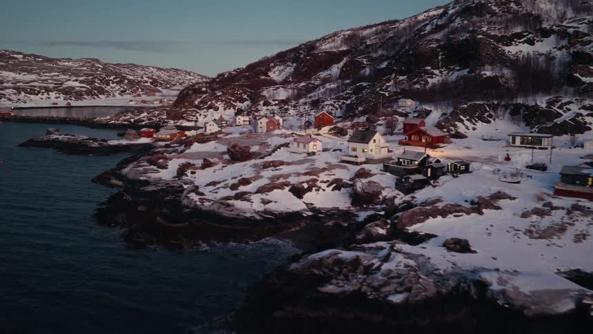 An aerial drone slowly flies towards a small, remote fishing village covered in snow along a rocky coastline during a peaceful winter twilight