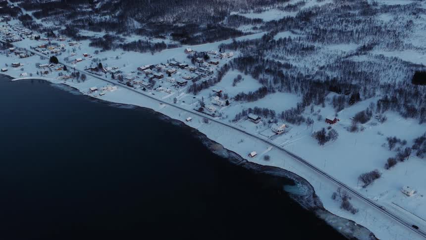 An aerial view reveals a tranquil, snow-covered coastal village and a lone car driving along the icy shoreline in the winter twilight
