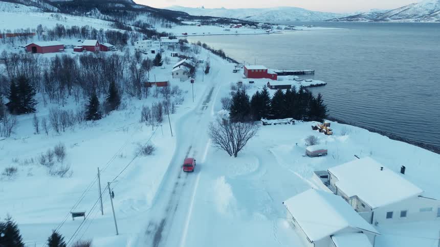An aerial shot follows a red car driving along a snow-covered road through a remote coastal village in Norway during winter