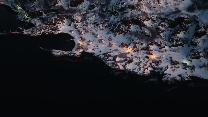 A top-down aerial view descends over a small, illuminated village nestled along a snowy coastline at night