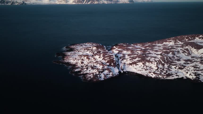 A slow aerial pullback reveals a snow-dusted rocky peninsula surrounded by a calm arctic sea, with majestic mountains in the distance