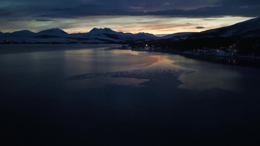 Slow aerial pullback reveals a breathtaking winter sunset over an icy fjord, with majestic snow-covered mountains in the distance