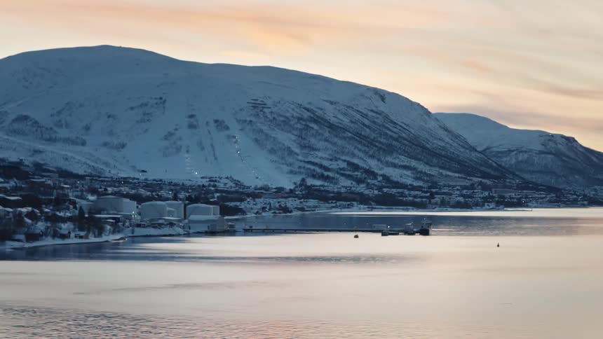 A tranquil panoramic view of a snow-covered coastal town nestled beside a calm fjord at the base of majestic mountains during sunrise