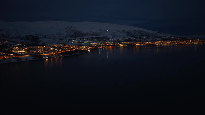 A tranquil night view of a glowing coastal town nestled against snow-covered mountains, its lights reflecting on the dark water