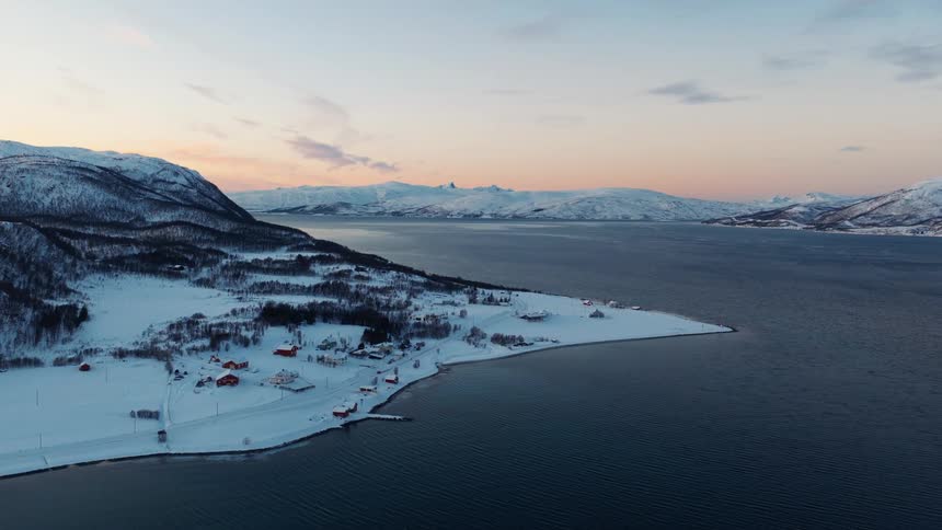 An aerial shot captures a tranquil, snow-covered village nestled on a peninsula by a fjord during a winter sunset in Norway