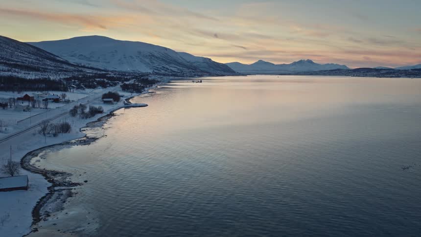 An expansive aerial view captures a serene Norwegian fjord at sunset, with snow-covered mountains and a quiet coastal road