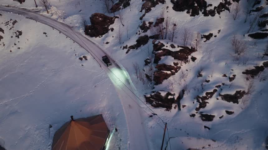 Aerial top-down view of a car driving along a snowy road into a remote Nordic village at dusk