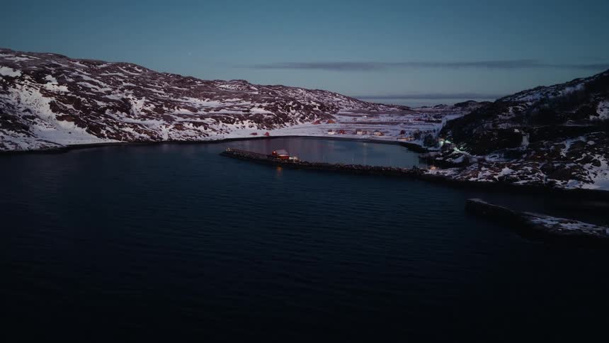 A slow aerial push-in shot reveals a tranquil, snow-covered coastal village nestled in a fjord during the quiet blue hour of twilight