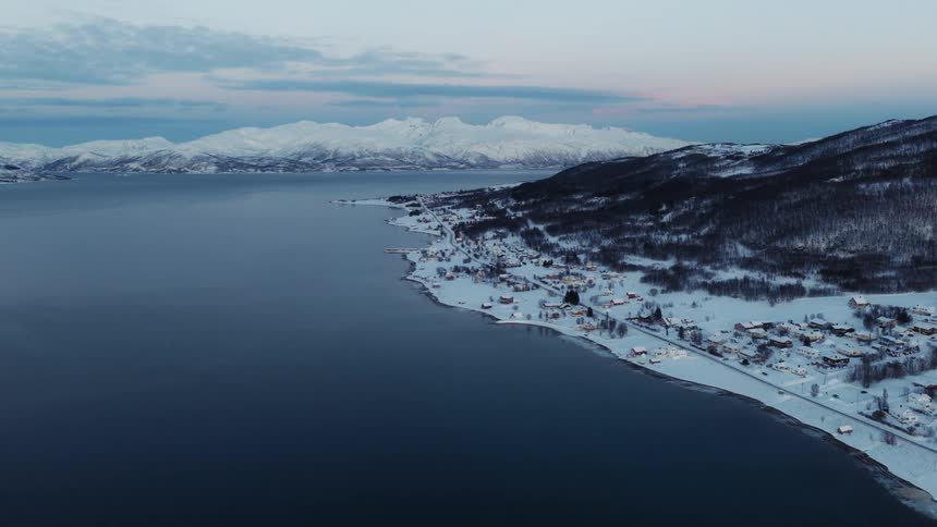 A stunning aerial shot of a tranquil, snow-covered coastal town in Norway with majestic mountains in the background at twilight