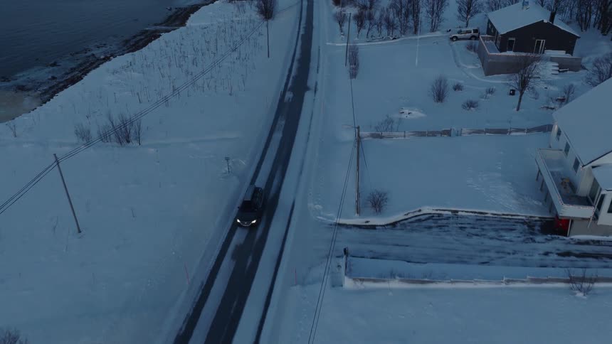 An aerial drone follows a car driving on a snowy road through a quiet coastal village at dusk
