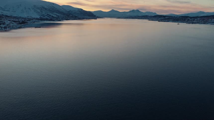 An aerial view tilts up from the dark, rippling water to reveal a vast, serene fjord surrounded by snowy mountains under a pastel sunset sky