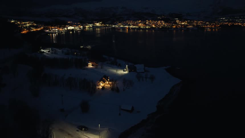An aerial view captures a cozy illuminated house on a snowy peninsula at night, with a distant town glowing across the water