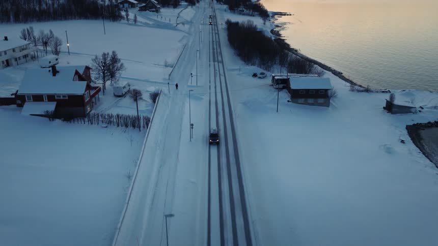 Cinematic aerial view of a car driving along a snow-covered road beside a fjord, revealing a vast winter mountain landscape at dusk