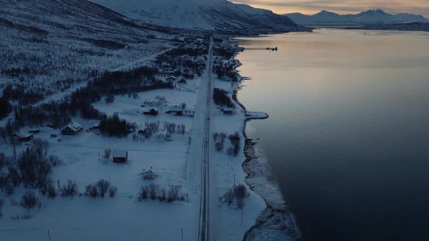 A serene aerial view of a snow-covered coastal village in Norway nestled between mountains and a fjord at sunset