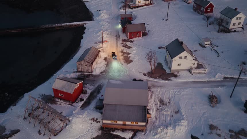 An aerial view follows a vehicle driving down a snowy road through a remote Norwegian fishing village at dusk