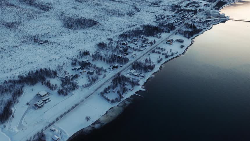 Drone pulls back over a tranquil, snow-covered coastal village nestled between a dark fjord and a wintery landscape at dusk