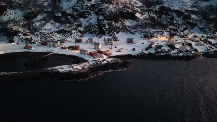 An aerial view captures the serene beauty of a small, illuminated fishing village nestled on a snowy coastline at twilight