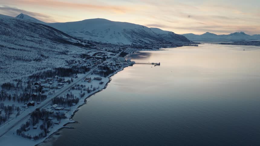 Static aerial view of a tranquil snow-covered Scandinavian village nestled between mountains and a calm fjord at dusk