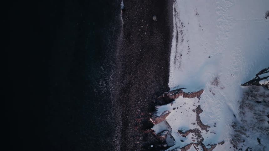 A mesmerizing top-down aerial view slowly rises over a remote, snowy coastline where the dark ocean meets a pebble beach