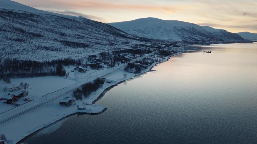 Aerial footage ascends over a calm fjord, revealing a snowy coastal village nestled at the base of mountains during a serene winter sunset