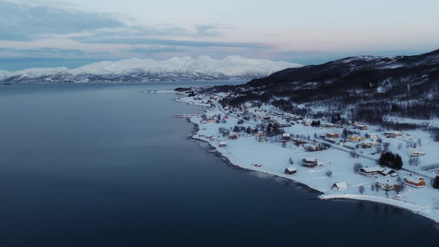 A serene aerial view captures a snowy coastal village nestled by a calm fjord at dusk with majestic snow-capped mountains in the background